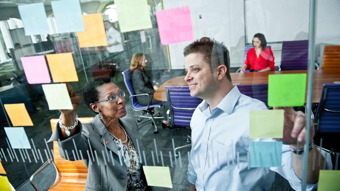 A white man and a black woman smiling behind a glass wall adorned with post-it notes.