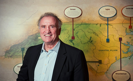 A middle-aged man, Walt Wolfram, stands in front of a map detailing the dialects of North Carolina.