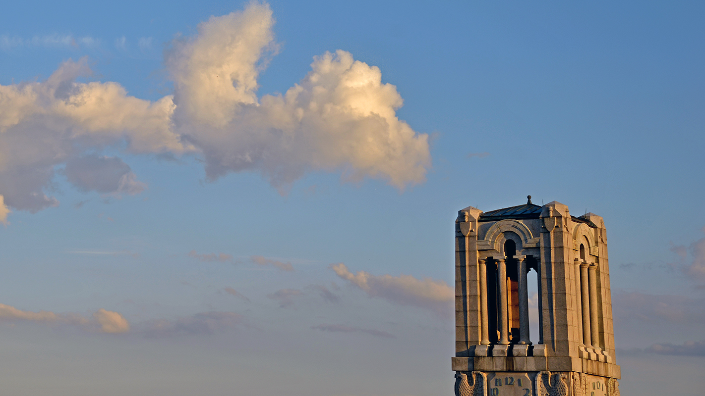 The top of the NC State Memorial Bell Tower peeks into view amid blue sky and clouds.