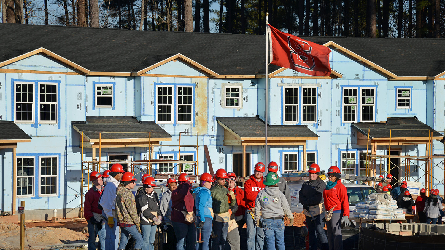 A group of Build-A-Block volunteers in hard hats gathered under an NC State flag.