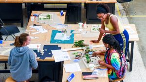 Three young women discuss a design project over a large desk.