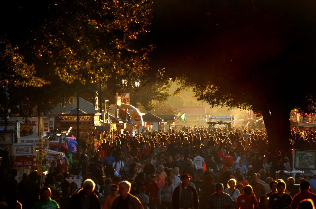 A huge crowd of fairgoers on the midway is illuminated by a warm autumn light.