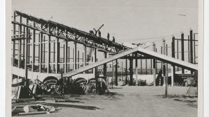 A black-and-white photograph shows construction workers climbing the steel skeleton of what will become Dorton Arena.