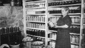 A woman looks carefully at a jar of preserves in this black-and-white image. Behind her are two shelves full of neatly-arranged jars.