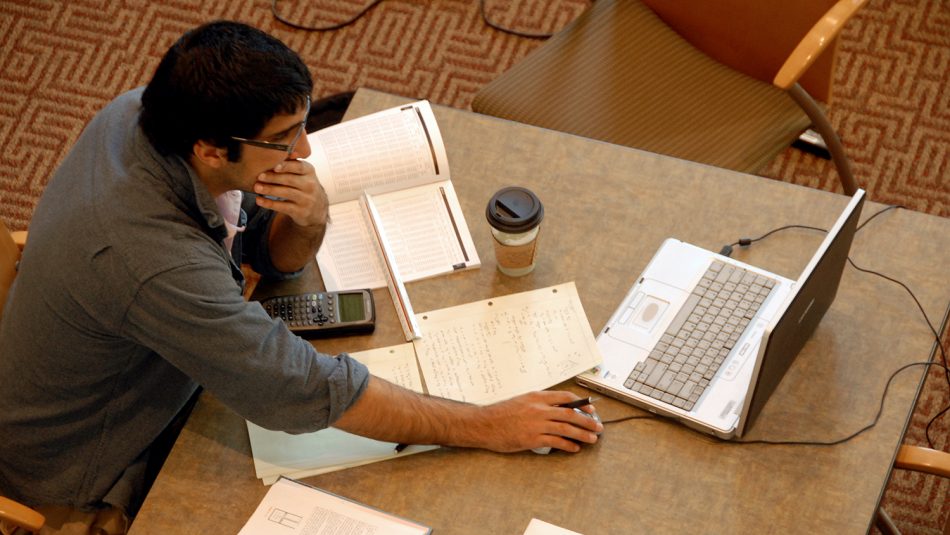 A student studies in the library with his laptop and books open, a cup of coffee at his side.