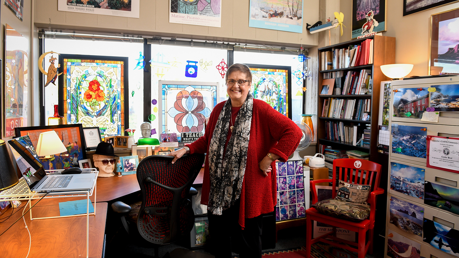 Candy Beal stands in her office before a large stained glass panel and many eclectic decorations.