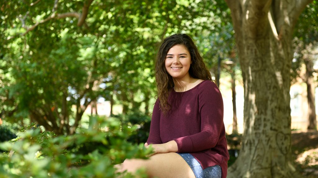 Incoming student Regan Mitchem sits on a bench surrounded by plants on NC State's main campus.
