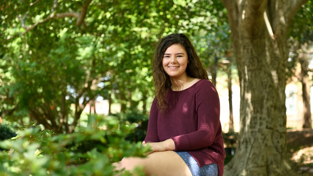 Incoming student Regan Mitchem sits on a bench surrounded by plants on NC State's main campus.