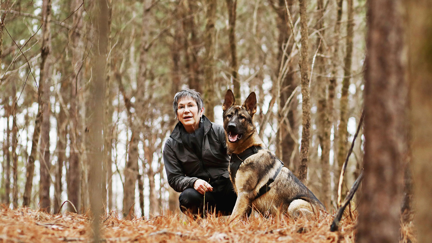 woman crouching down beside a seated German shepherd dog in the woods