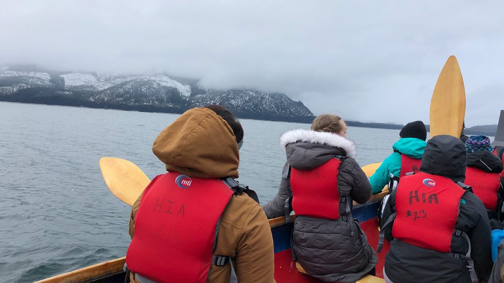 Students wearing heavy coats and lifejackets canoe across water off the coast of Alaska, with snowy mountains in the distance