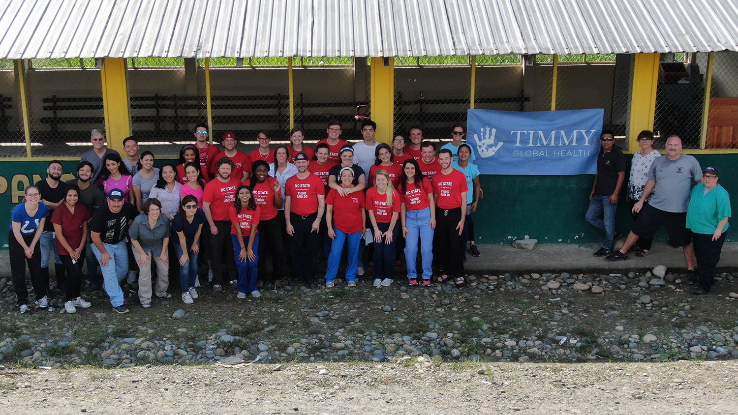 A team of students and leaders wearing NC State shirts stand outside a rural clinic in Ecuador with a TImmy Global Health banner