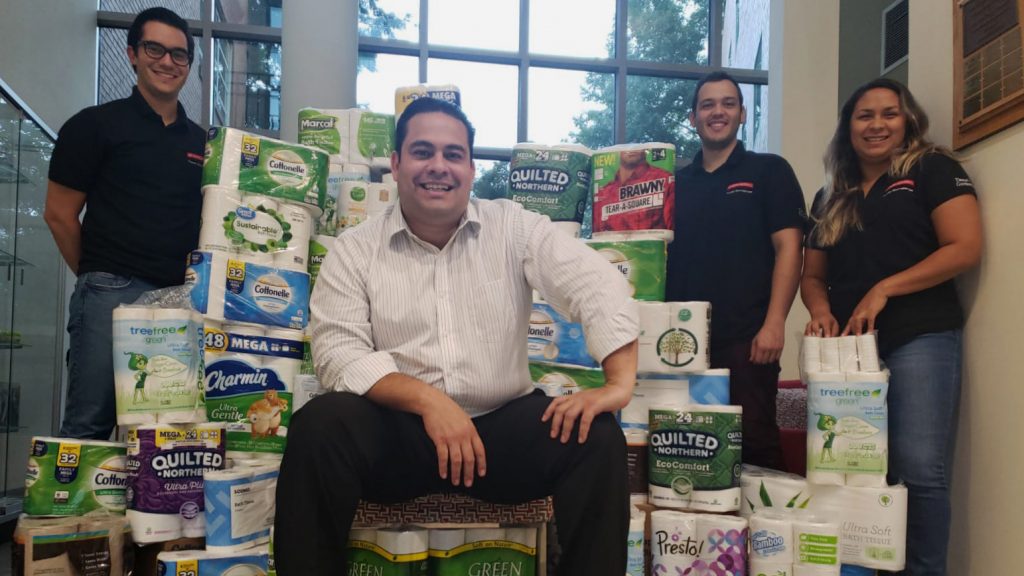 Ronalds Gonzalez sits on a make-shift chair made out of toilet paper packages while posing with three students.