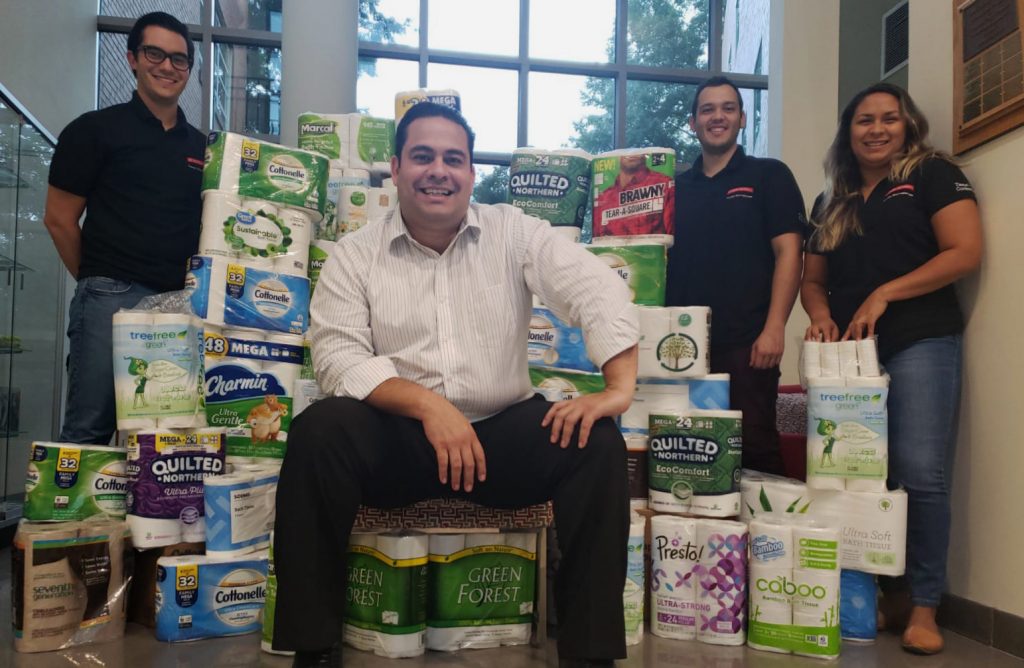 Ronalds Gonzalez sits on a make-shift chair made out of toilet paper packages while posing with three students.