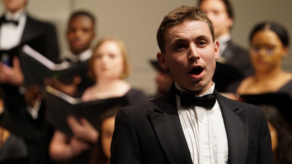 A young man sings on stage with a choir behind him.