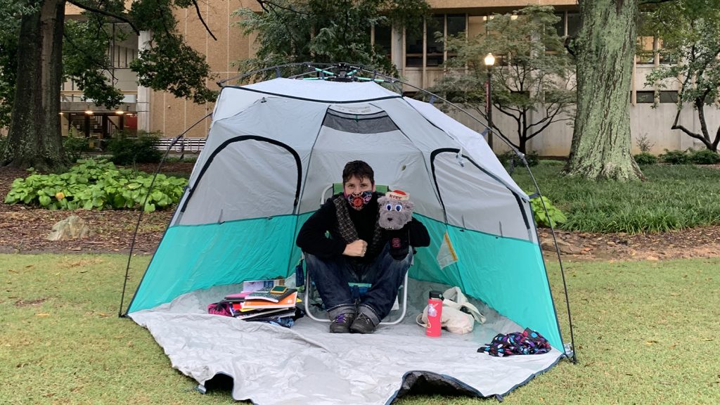 Valerie Faulkner sits in a tent, wearing a face mask and holding a Mr. Wuf puppet, while she waits for students to pick up their journals.