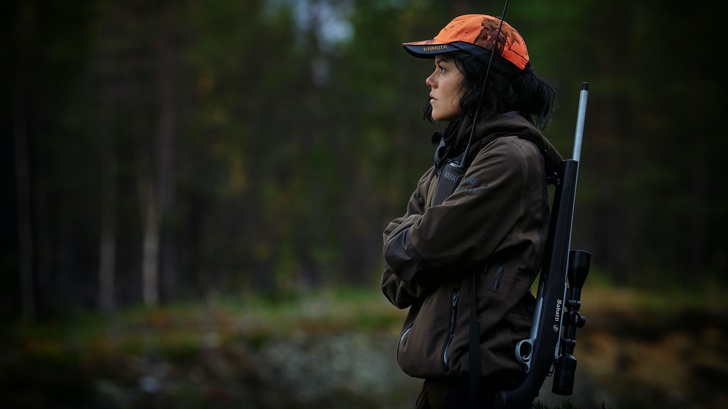 woman in hunting attire stands with rifle