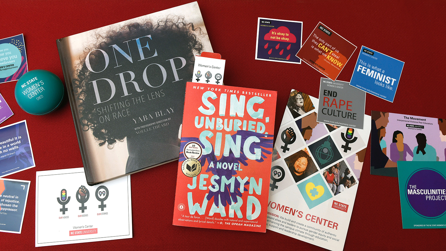 A flat lay image that includes Women's Center stickers, flyers and a copy of the book Sing, Unburied, Sing.