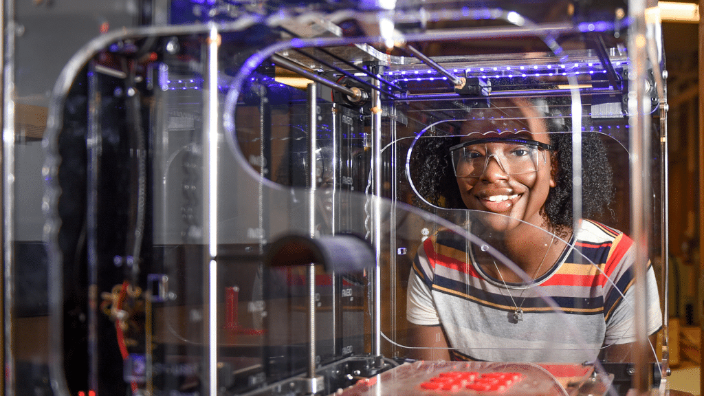 A young woman stands behind a 3D printer.