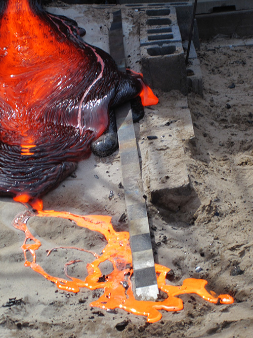 Metallic lava forms a volcano-like cooling peak while orange lava flows out of it onto dirt