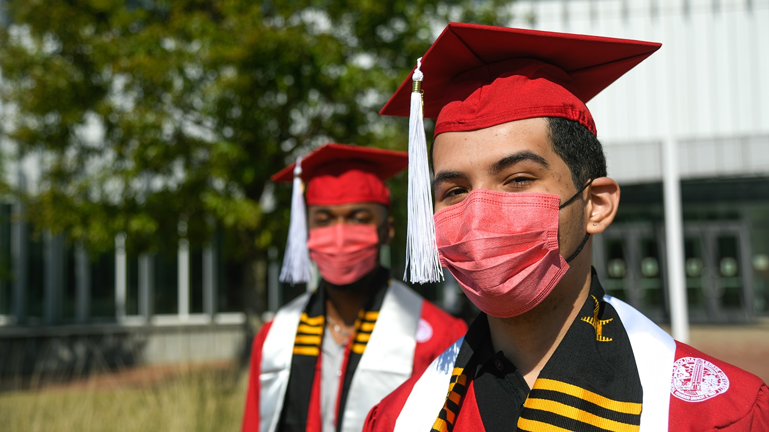 Jeancarlo Ferman, in the foreground, and Gabriel Montague, in the background, wear their red commencement robes and mortar boards, stoles and red face masks outside. 
