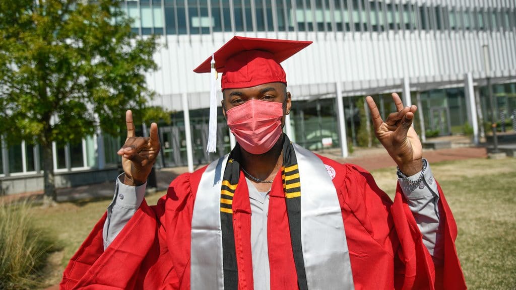 Gabriel Montague makes wolf hands outside while wearing his red undergraduate regalia and a matching red face mask. 