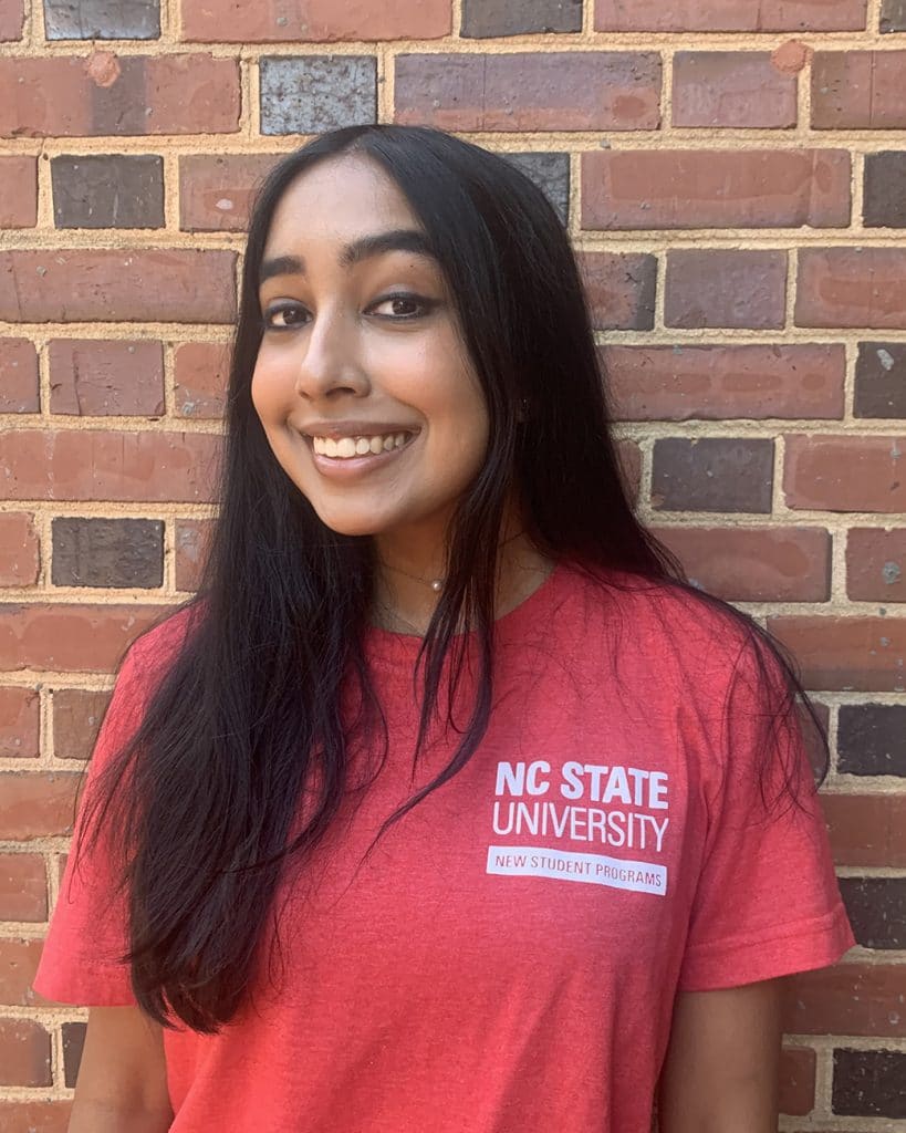 Esha Bhatnagar, wearing a red NC State t-shirt, stands for a portrait in front of a brick wall.