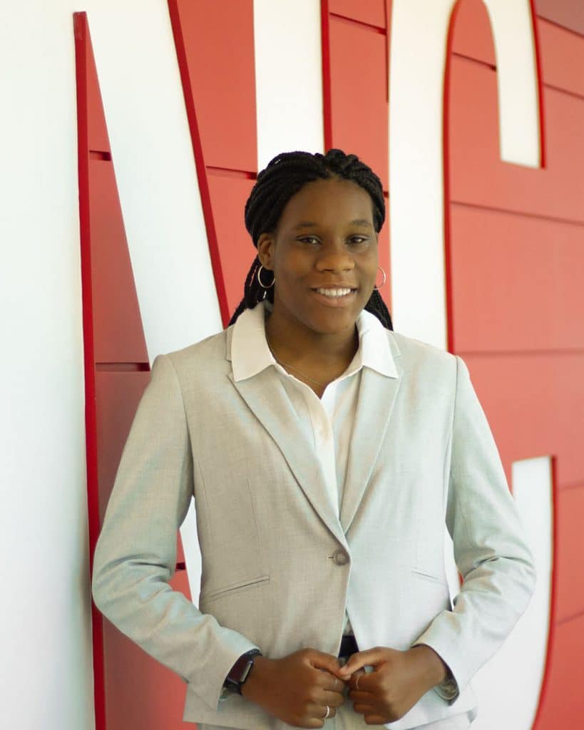 Kayla Alford, wearing a cream jacket, stands for a portrait in front of a red and white background.