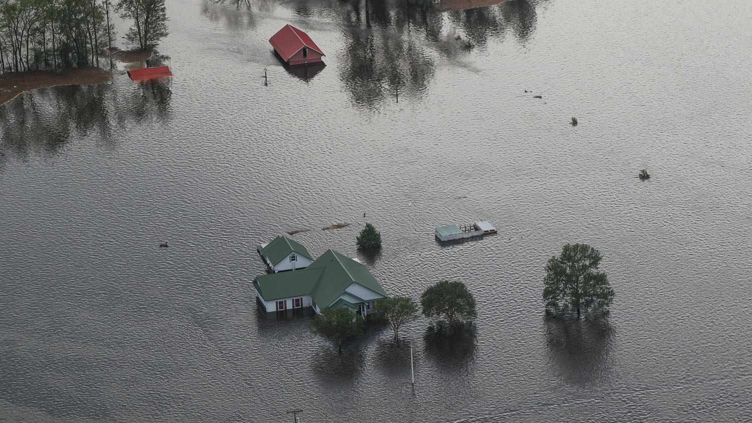 aerial view of a farmhouse, vehicles and outbuildings during a flood. all of the structures are submerged, leaving only roofs exposed