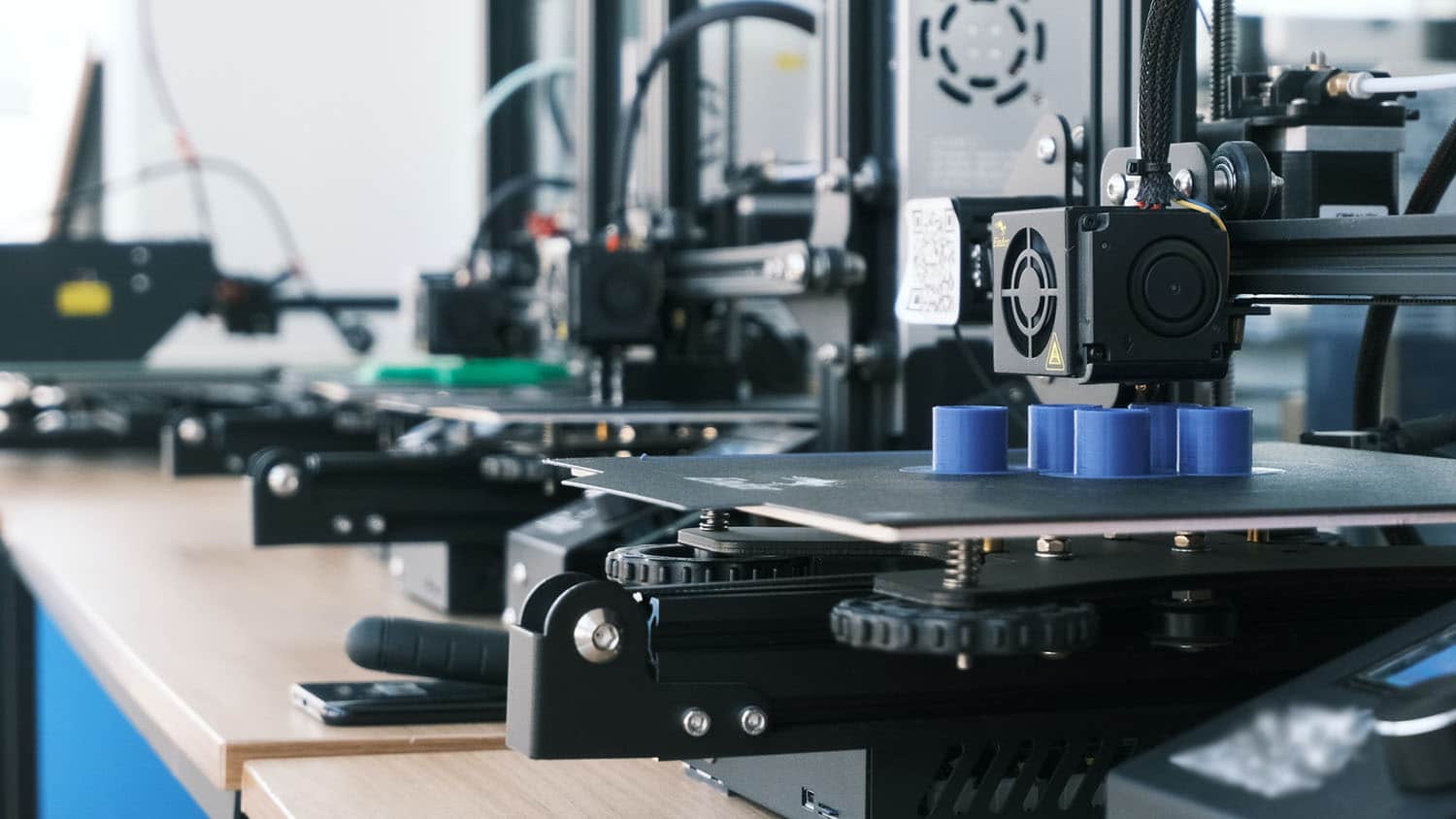 several 3d printers are lined up on a workbench; one of them is printing an object