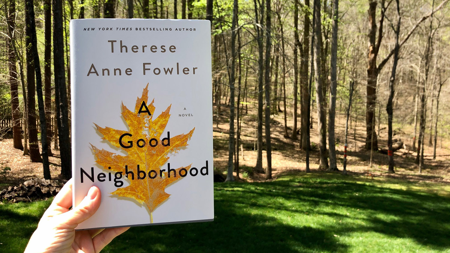 A hand holds Therese Anne Fowler's book "A Good Neighborhood" outside in a wooded area.