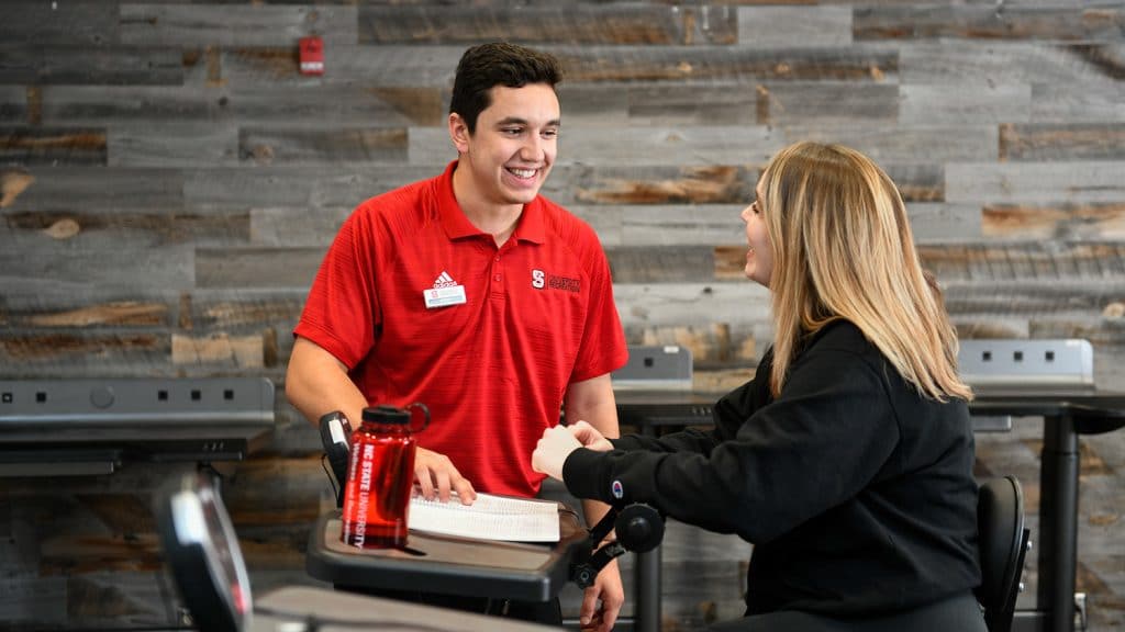 Man in a red NC State shirt talks with another student sitting on a stationary bike