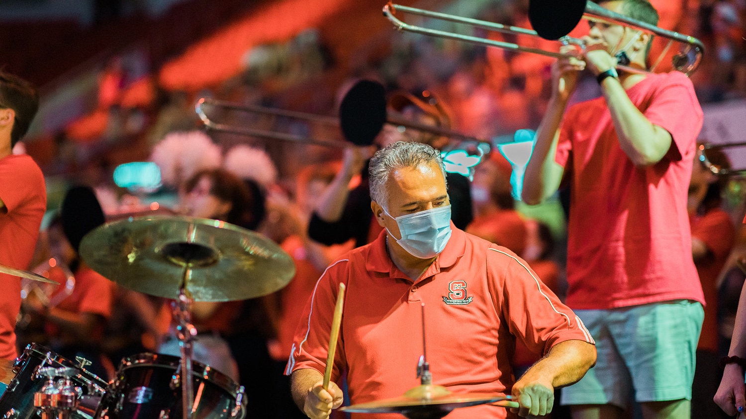A man in a red shirt with the NC State logo plays the drums. Another man in a red shirt plays the trombone. 