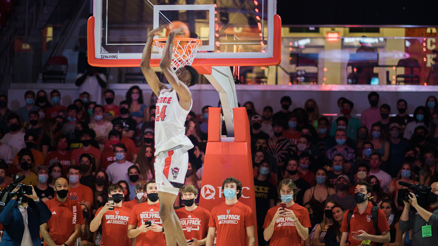 An NC State basketball player dunks a basketball into a net in front of a crowd of fans. 