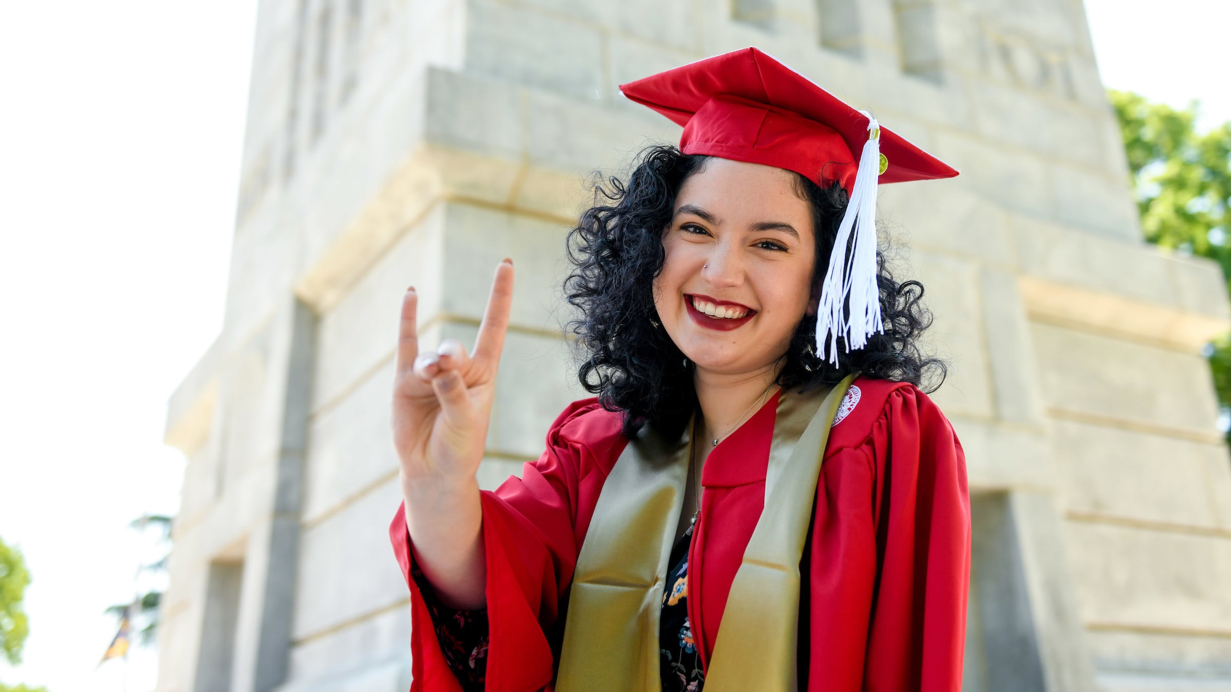 A student in a red cap and gown makes the Wolfpack sign in front of the NC State Belltower.