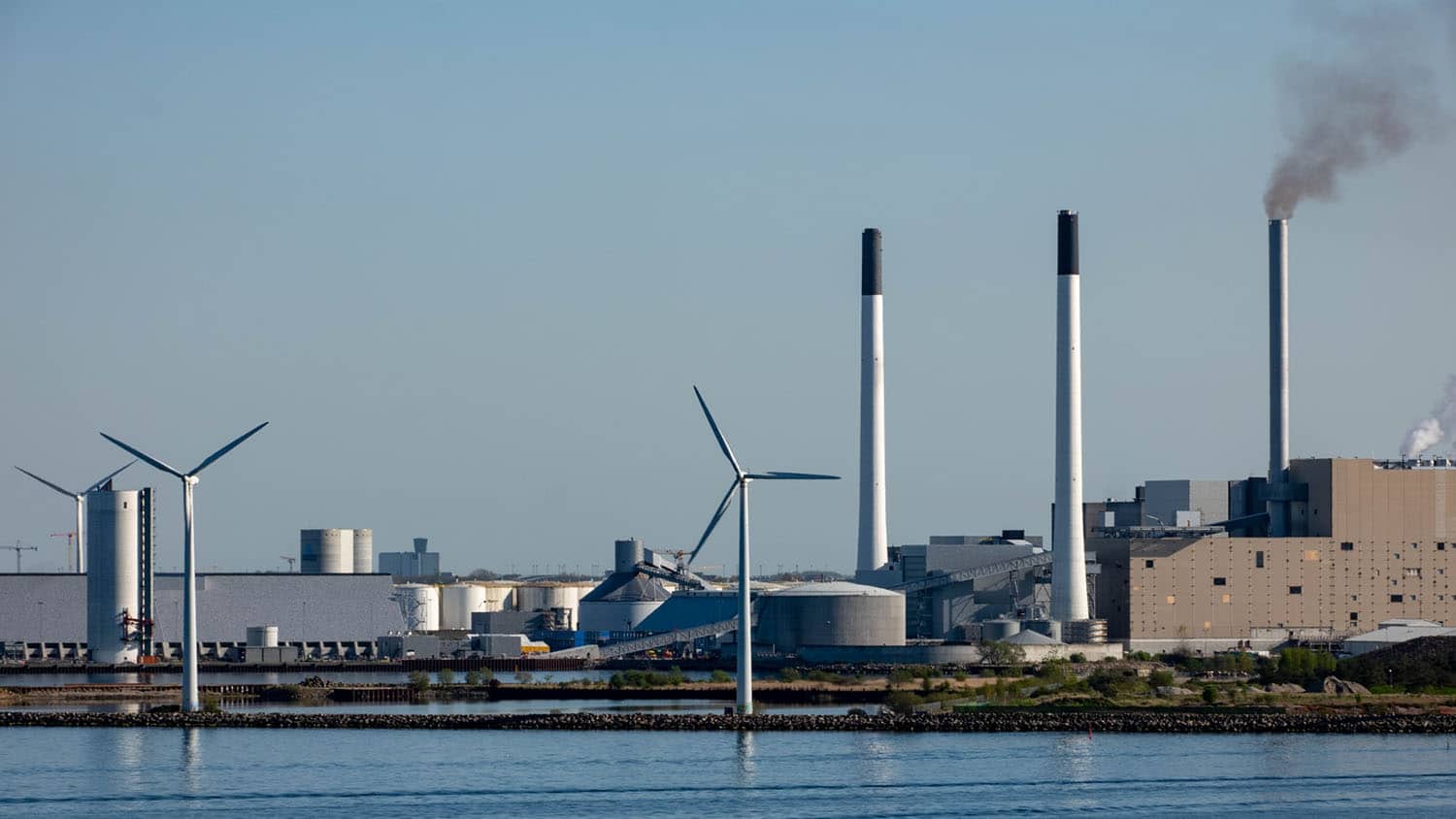 Photo looks out across the water at a series of windmills and the smokestacks of a power plant