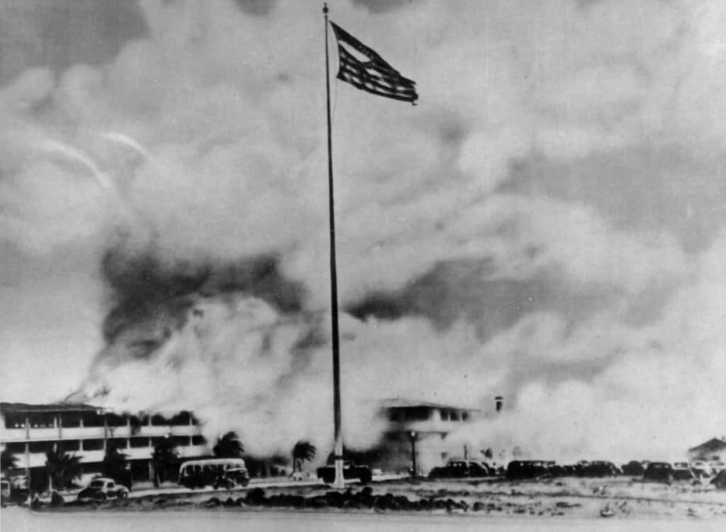 View of the burning barracks at Hickam Field and the shredded U.S. flag on the flagpole after the Japanese attack on Pearl Harbor. Photo: National Park Service.