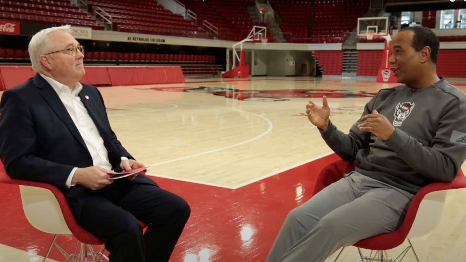 Randy Woodson and Kevin Keatts talk while sitting on a basketball court.