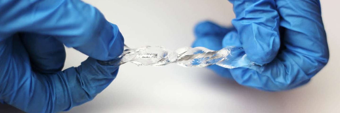 A person wearing blue surgical gloves twists a clear soft material to show its flexibility