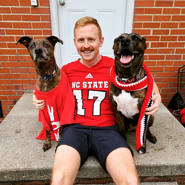 man wearing NC State football shirt posing with two dogs
