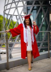Anna Jump, wearing a white dress and her red commencement attire, makes the wolf symbol with her hand outside of Talley Student Union.
