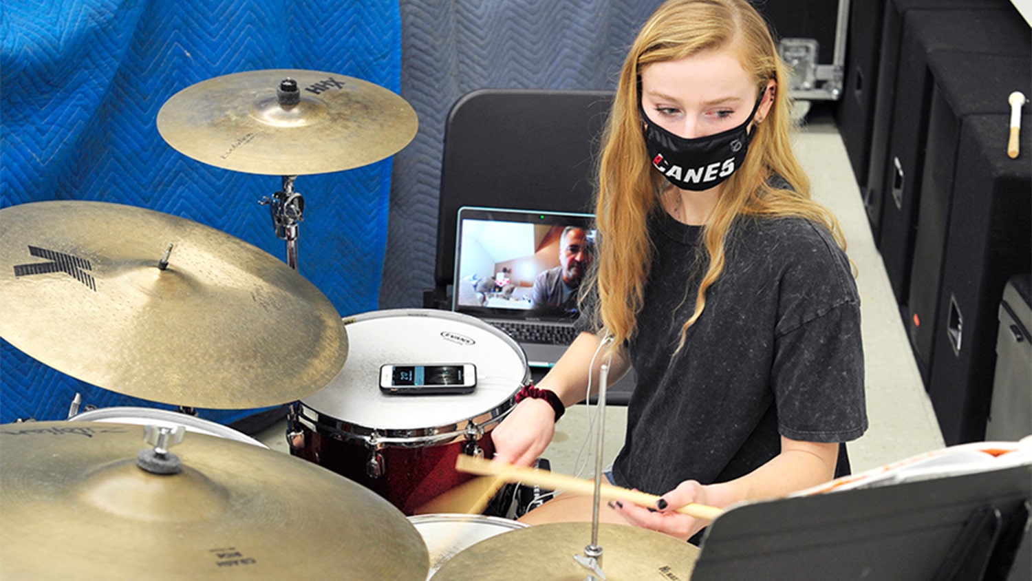 AnnE Ford practices playing the drums while wearing a black face mask. Her phone is sitting on one of her drums, and a video call with her professor is visible on a laptop screen behind her. 