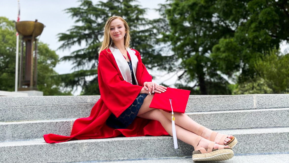AnnE Ford sits on the steps of Memorial Belltower, wearing her red graduation gown and holding her mortar board.