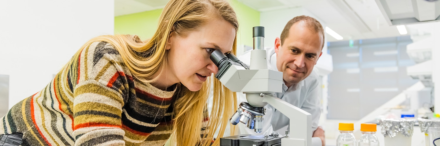 Postdoctoral researcher Lisa Van den Broeck looks into a microscope while STEPS Center director Jacob Jones watches on by her side..