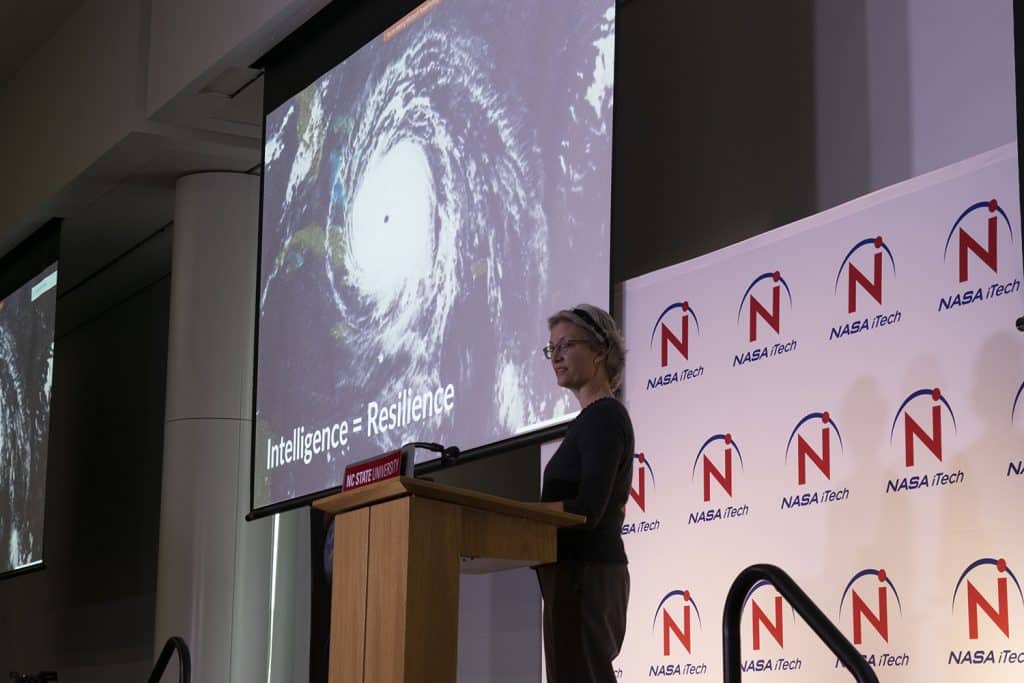 Warrillow stands at a lectern in front of a screen that displays a hurricane formation.
