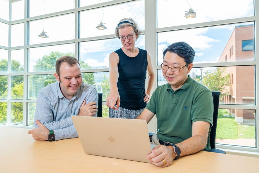 Zambon, Warrillow and He sit at a table inside and look at a laptop. They sit in front of a window which overlooks Centennial Campus.