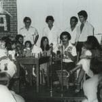 A guitar player sits at a microphone, surrounded by a group of adults and small children, during an international fair in 1980, in this black and white archival photo.