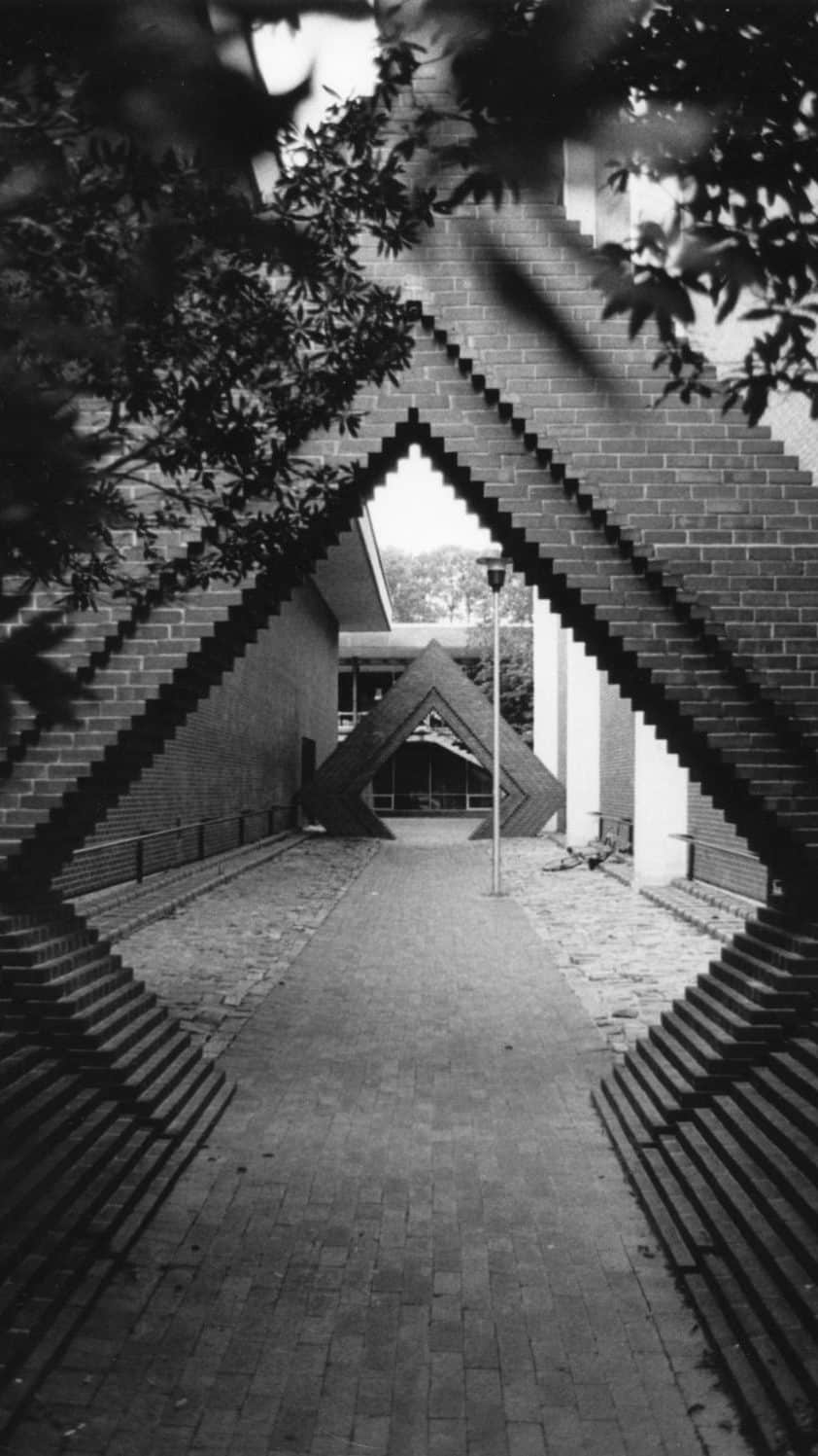 A black and white archival photo shows a view of a brick-paved walkway between the original University Student Center and Price Music Center.