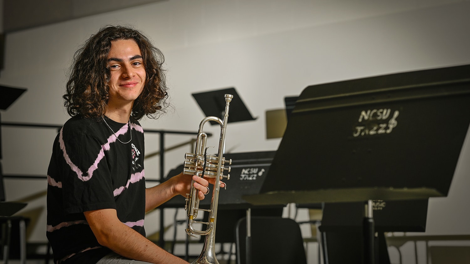 Aidan Dumain holds a trumpet in a rehearsal room, surrounded by music stands that say "NCSU Jazz".