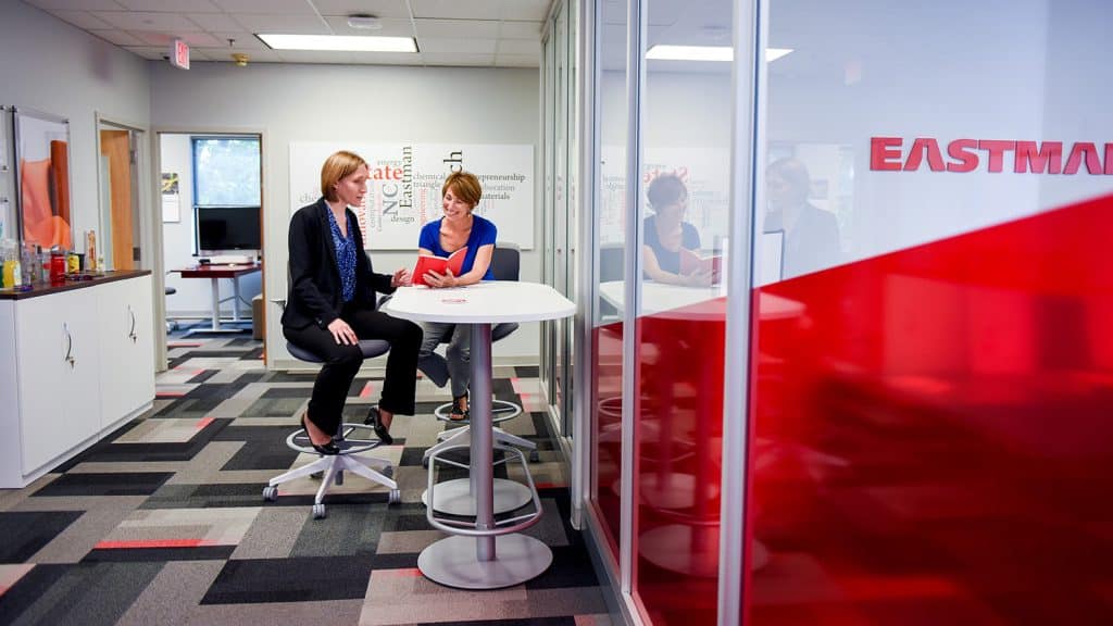 Two women sit at a round table at Eastman’s innovation center on Centennial Campus. They are looking at through a red notebook.
