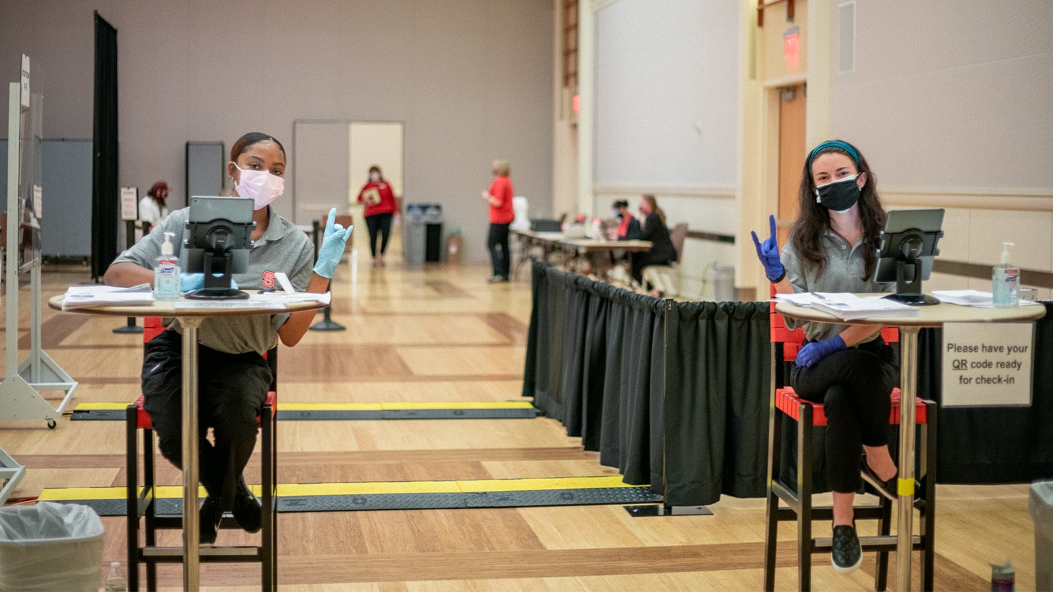 Two employees sit at check-in tables and make wolfie signs at a vaccination clinic